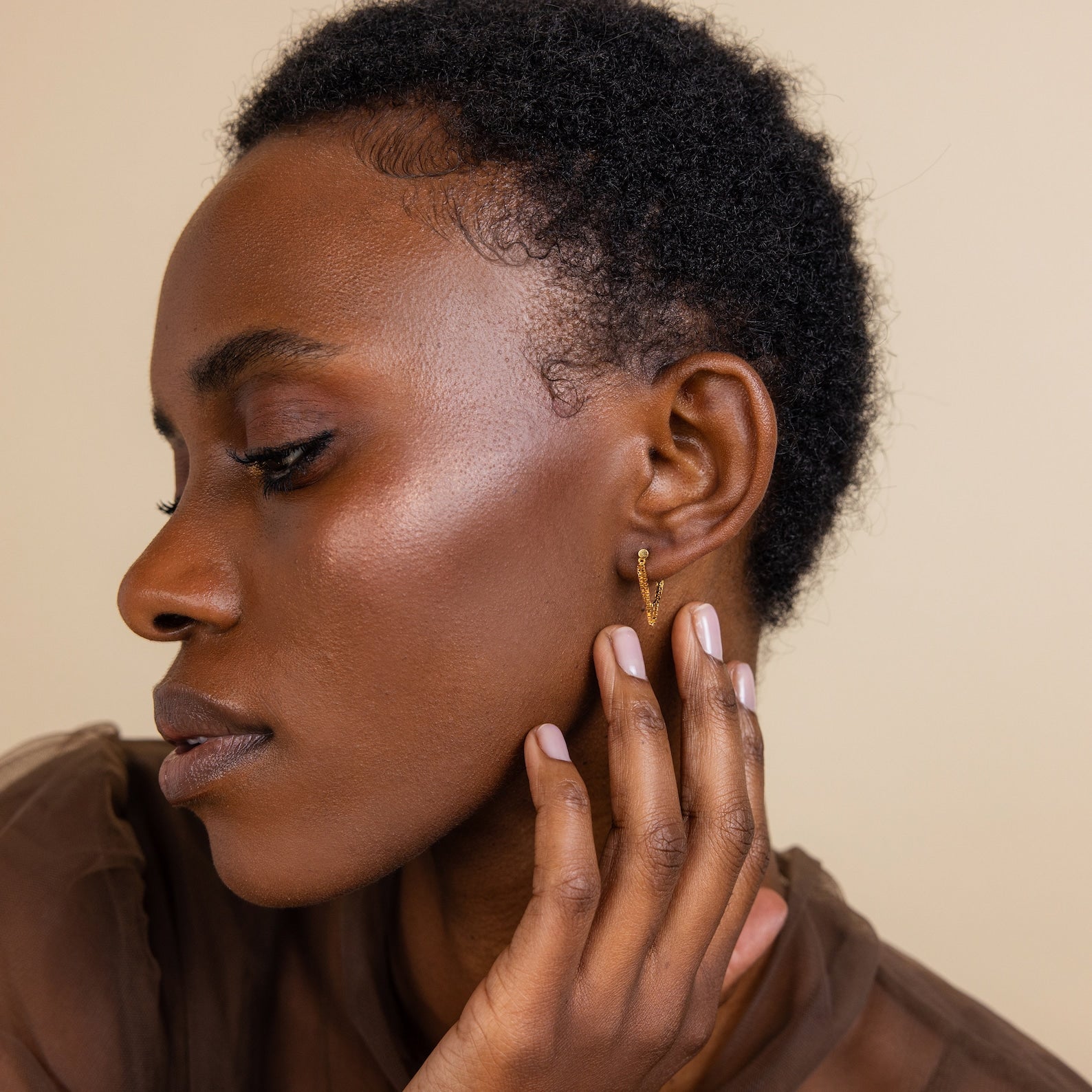 A woman with short, natural hair touches her face, highlighting Flatback Chain Earrings from her minimalist stack and dewy makeup in side profile.