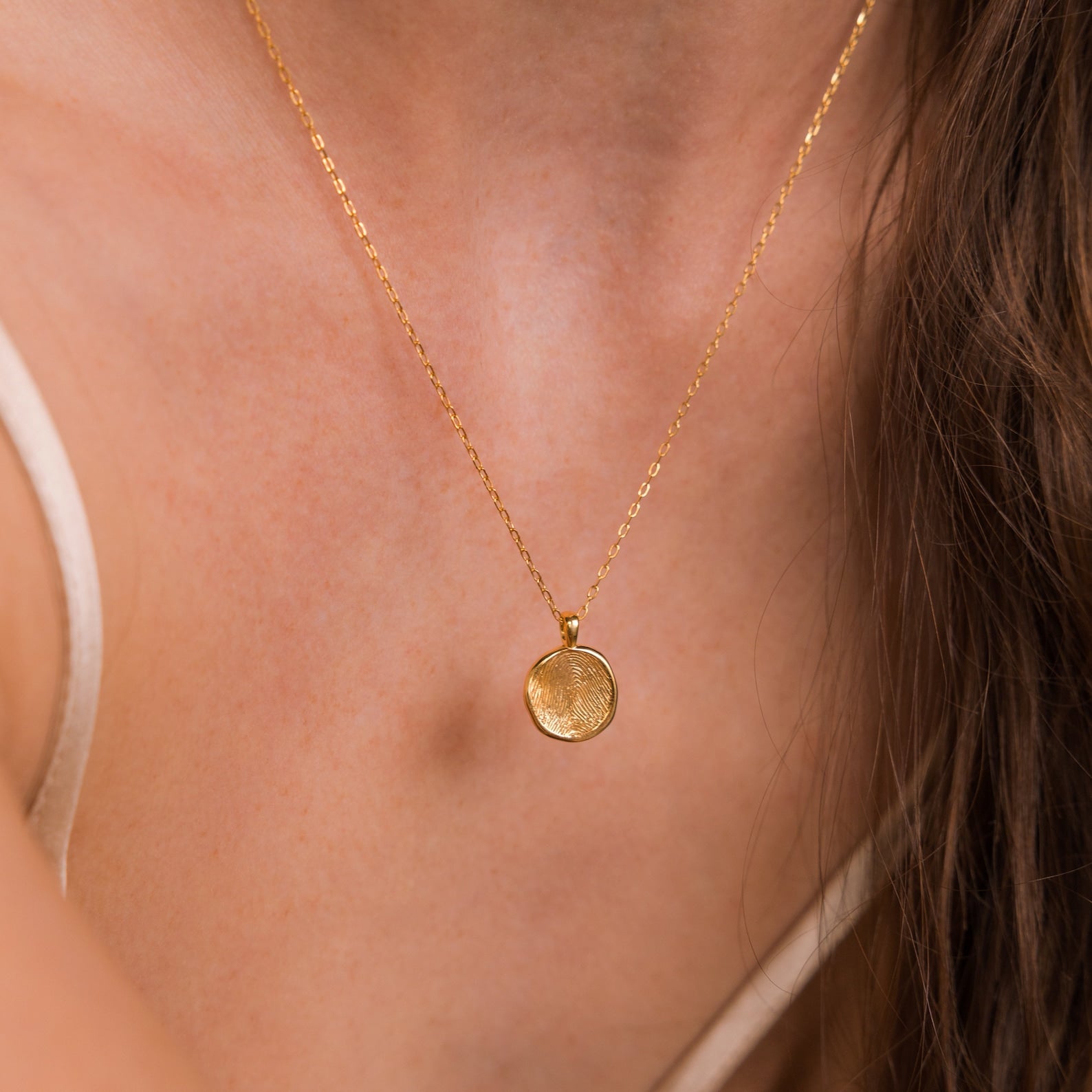 The Fingerprint Coin Necklace, featuring a round gold pendant, is worn with a beige top—a heartfelt choice for Mother’s Day.