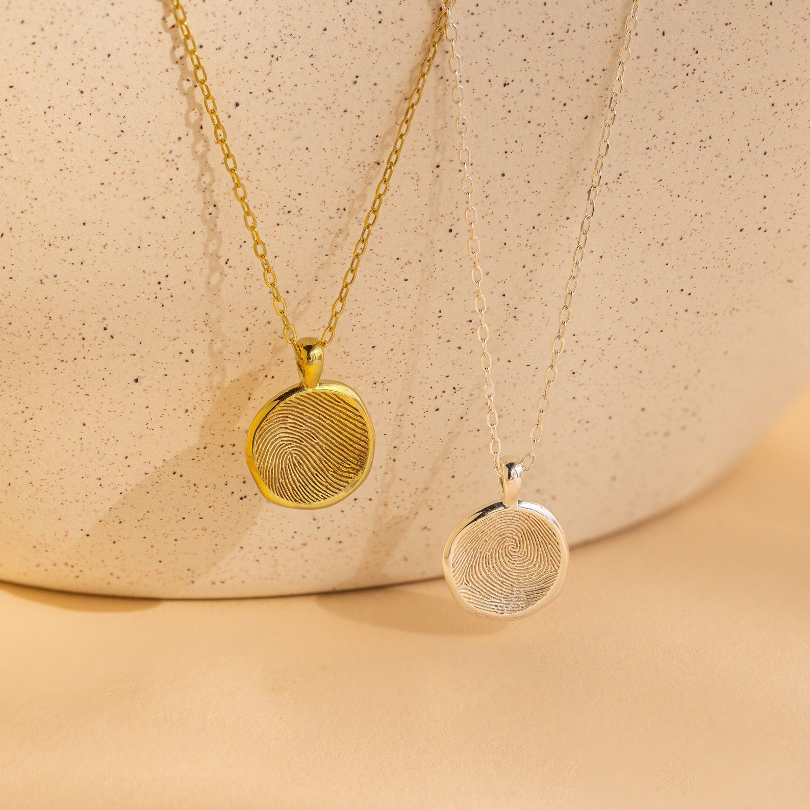 The Fingerprint Coin Necklace features a round pendant in gold and silver, displayed on a textured beige background—an elegant piece ideal as a Mother's Day gift.