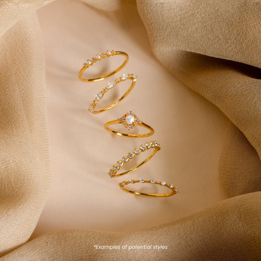 Five Mystery Rings in gold with small gemstones, displayed on beige fabric. These unique pieces are part of our eclectic jewelry collection. Text below reads: Examples of potential styles.