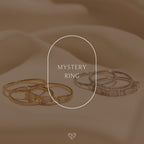 Two sets of gold and silver Mystery Rings from our unique jewelry collection rest on soft beige fabric, with "Mystery Ring" displayed in the center.