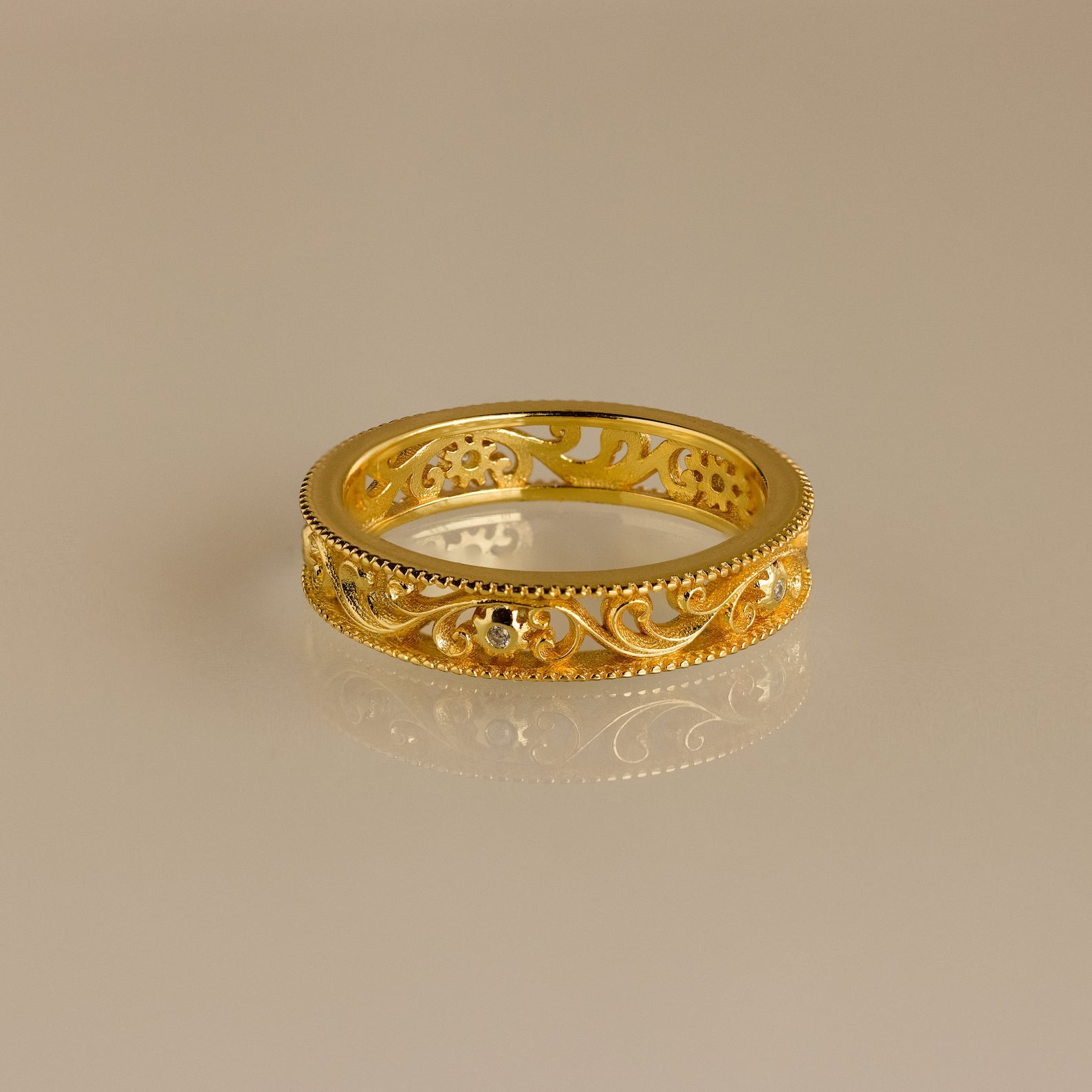 The Filigree Flower Ring is a vintage-inspired gold band featuring ornate filigree and delicate floral patterns, softly reflecting on a neutral surface.