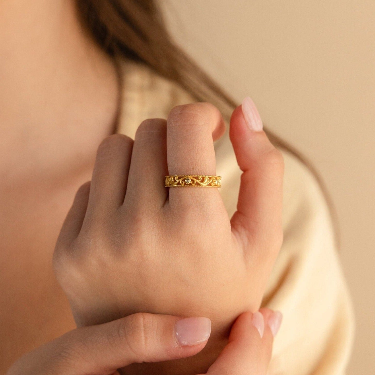 A hand wears the Filigree Flower Ring, showcasing its ornate gold band with intricate floral details, as fingers gently touch the elegant vintage-inspired design.
