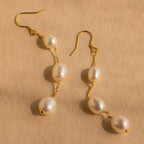Close-up of a pair of long gold pearl chain earrings set against a tan background.