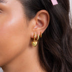 A woman with brown hair and a pink hair clip wears three hoop earrings, including Puff Heart Charm Huggies in Sterling Silver.