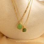 Two sophisticated Mystery Necklaces with green stone pendants rest on a textured beige surface, adding a touch of intrigue to any jewelry collection.