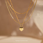 The Mystery Necklace features three sophisticated layers, including a heart pendant, displayed on a soft beige background—perfect for adding an elegant touch to your look.