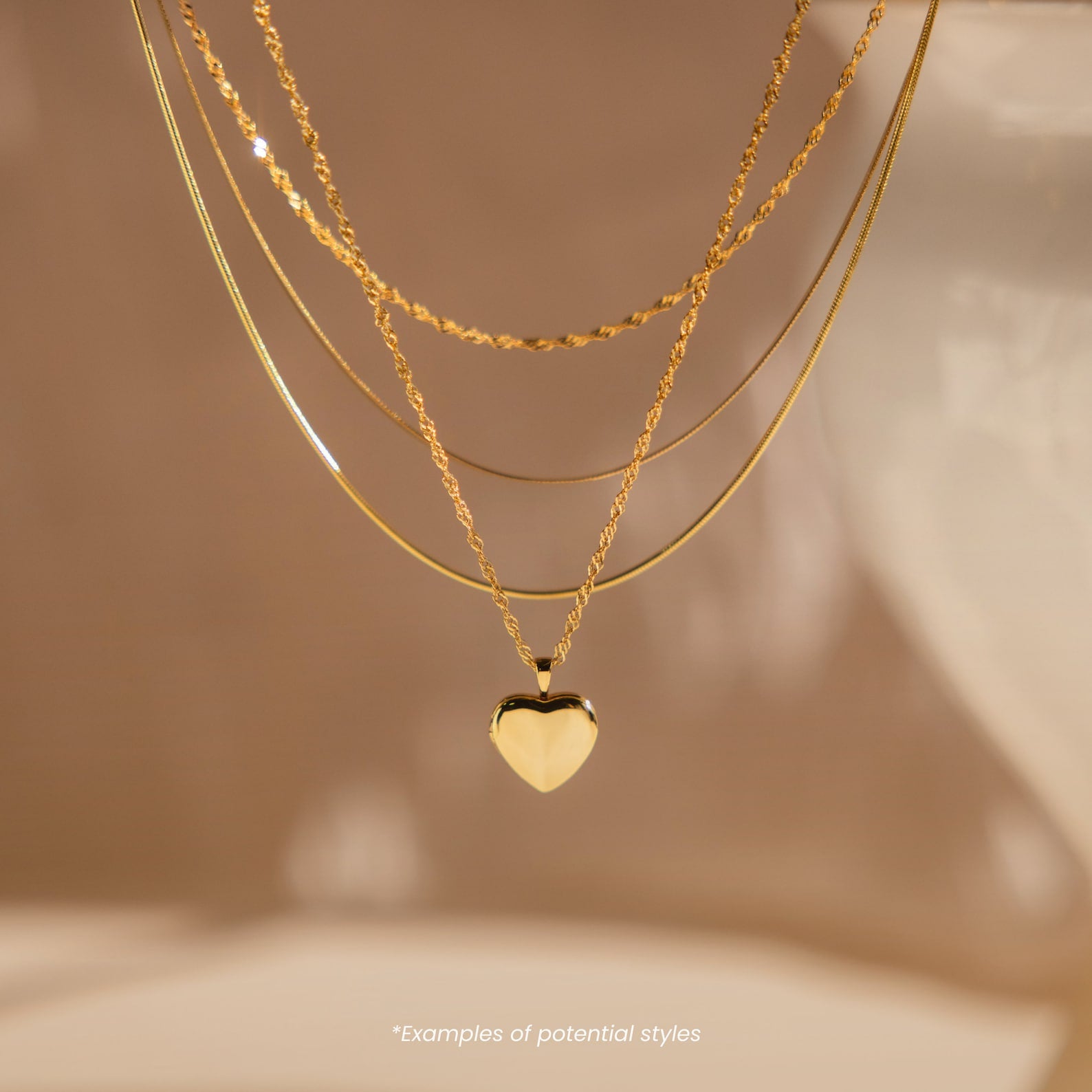 The Mystery Necklace features three sophisticated layers, including a heart pendant, displayed on a soft beige background—perfect for adding an elegant touch to your look.