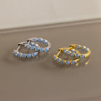 Two pairs of Inside Out Ivy Huggies—one in silver, one in gold—each set with delicate light blue gemstones, are elegantly displayed on a reflective surface.