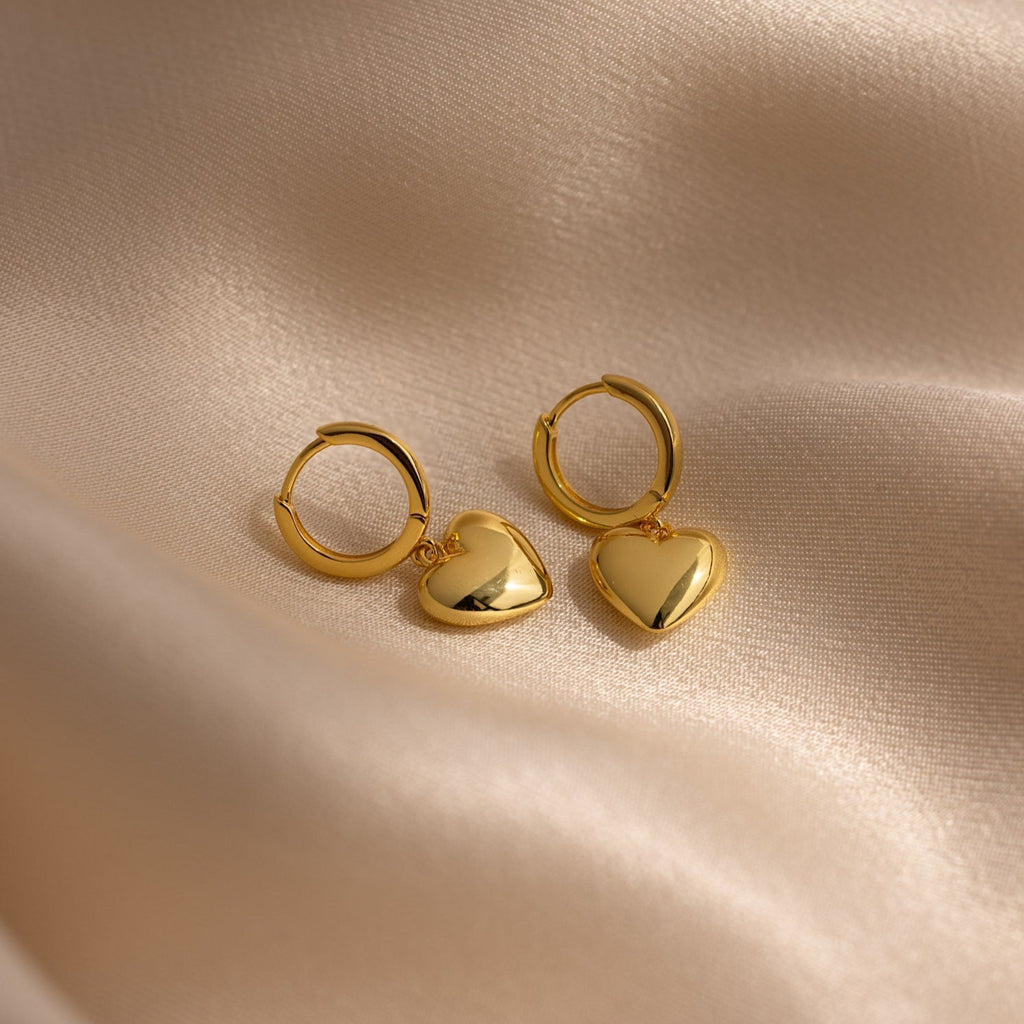 Puff Heart Charm Huggies are displayed on a soft, beige satin fabric background.