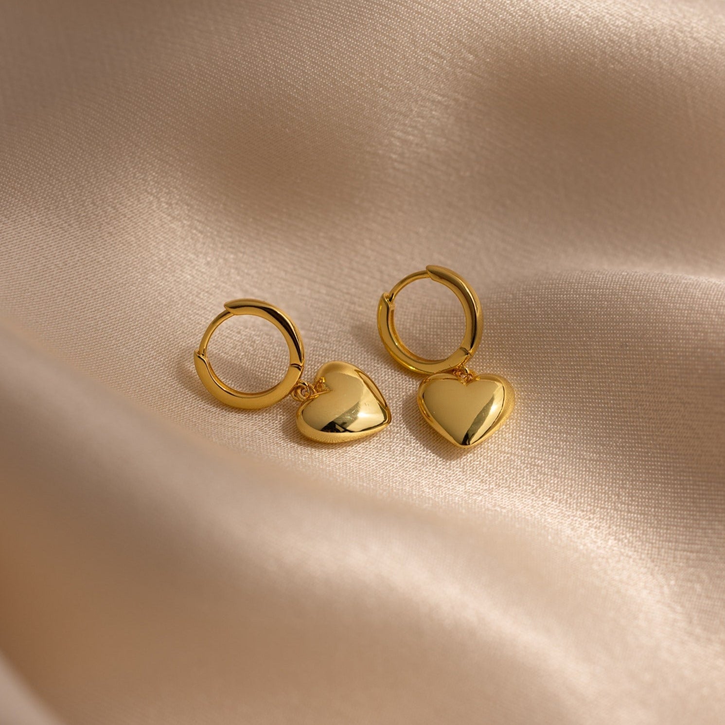 Puff Heart Charm Huggies are displayed on a soft, beige satin fabric background.