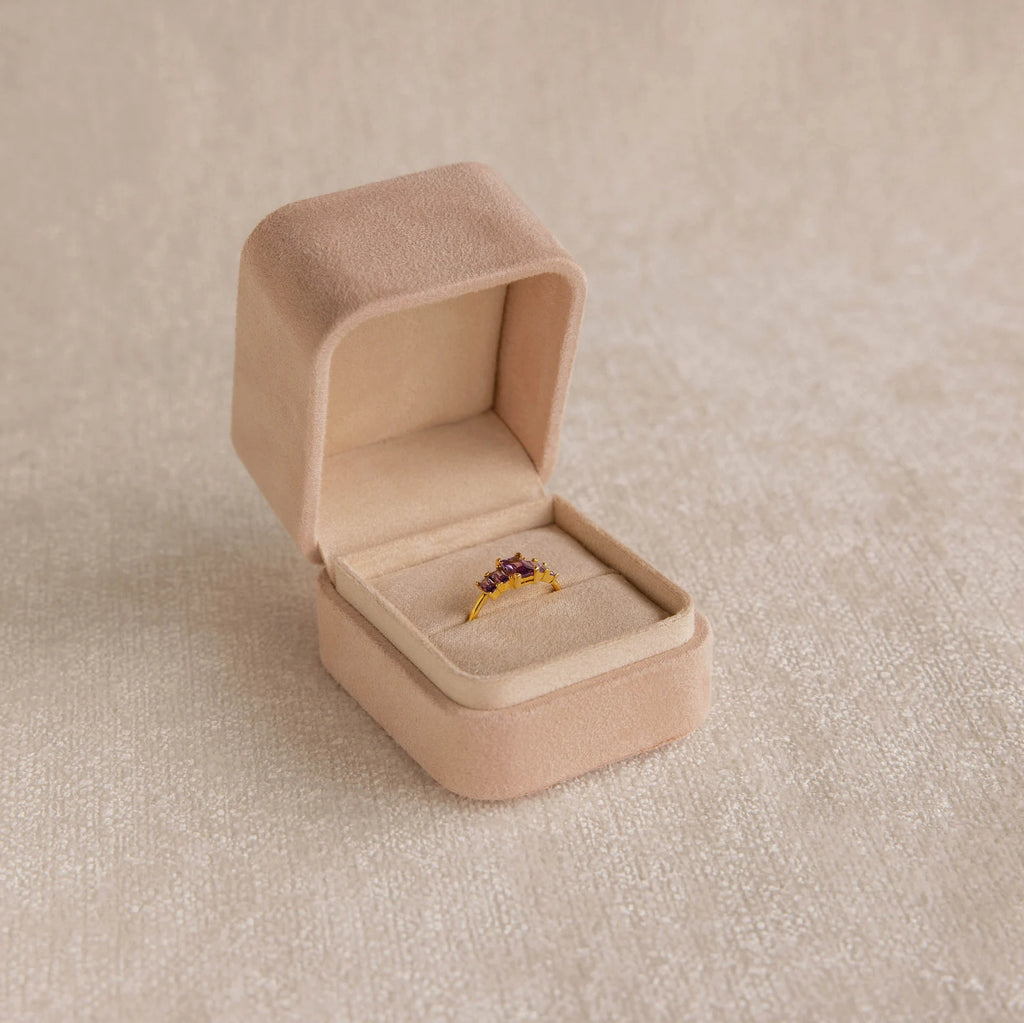 The Pink Suede Ring Box opens to reveal a gold ring with red gemstones, displayed on a neutral textured surface and highlighting its minimalist design.