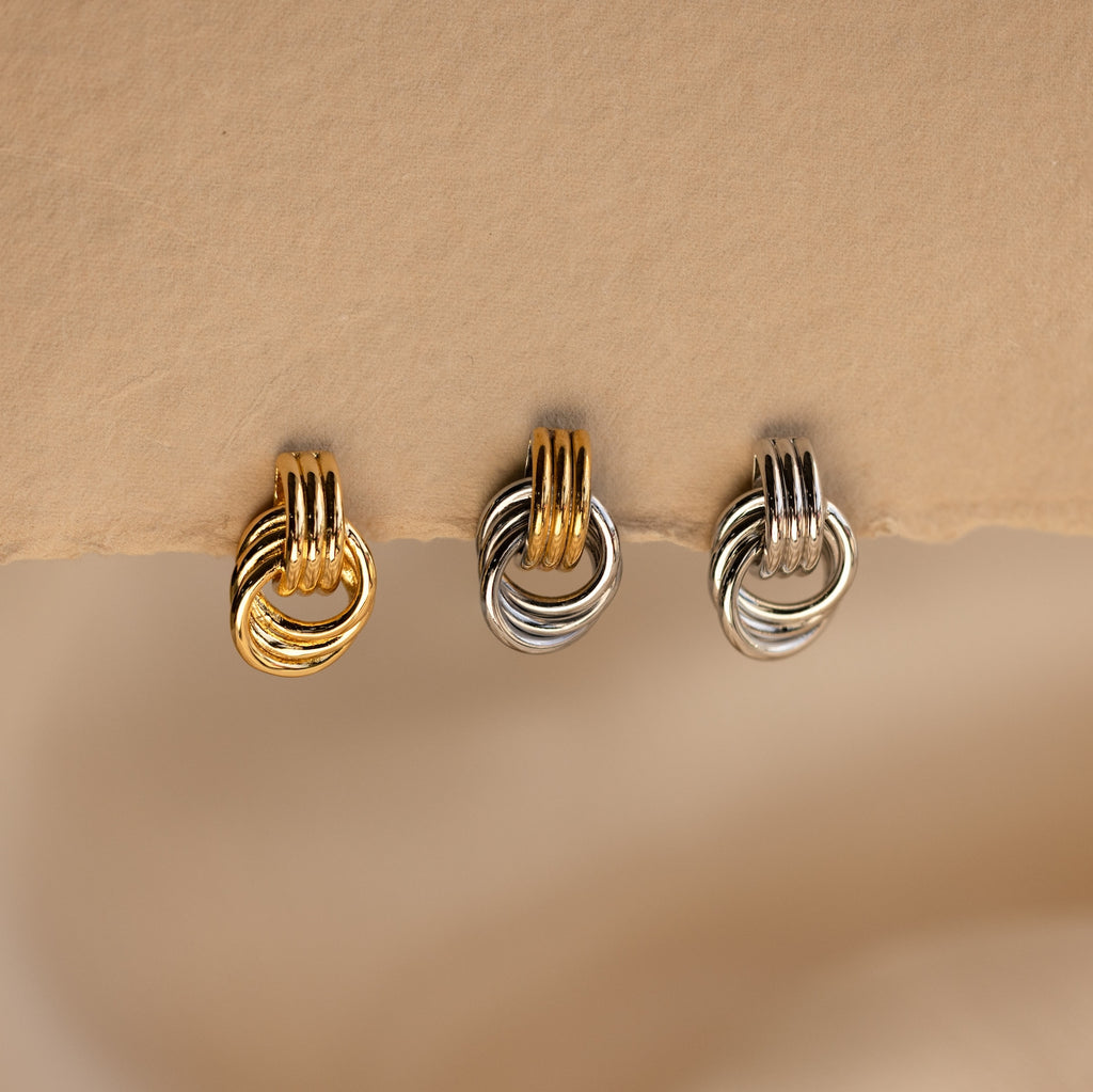 A close-up product shot of three individual Mini Triple Knot Earrings arranged in a horizontal row, suspended on the edge of a textured, natural-toned card. Each earring features three interlocking, high-polish bands forming a compact, elegant knot shape. The left earring is fully gold-toned, the center earring combines gold and silver tones for a mixed metal look, and the right earring is entirely silver-toned. 