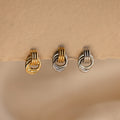 A close-up product shot of three individual Mini Triple Knot Earrings arranged in a horizontal row, suspended on the edge of a textured, natural-toned card. Each earring features three interlocking, high-polish bands forming a compact, elegant knot shape. The left earring is fully gold-toned, the center earring combines gold and silver tones for a mixed metal look, and the right earring is entirely silver-toned. 