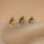 A close-up product shot of three individual Mini Triple Knot Earrings arranged in a horizontal row, suspended on the edge of a textured, natural-toned card. Each earring features three interlocking, high-polish bands forming a compact, elegant knot shape. The left earring is fully gold-toned, the center earring combines gold and silver tones for a mixed metal look, and the right earring is entirely silver-toned. 