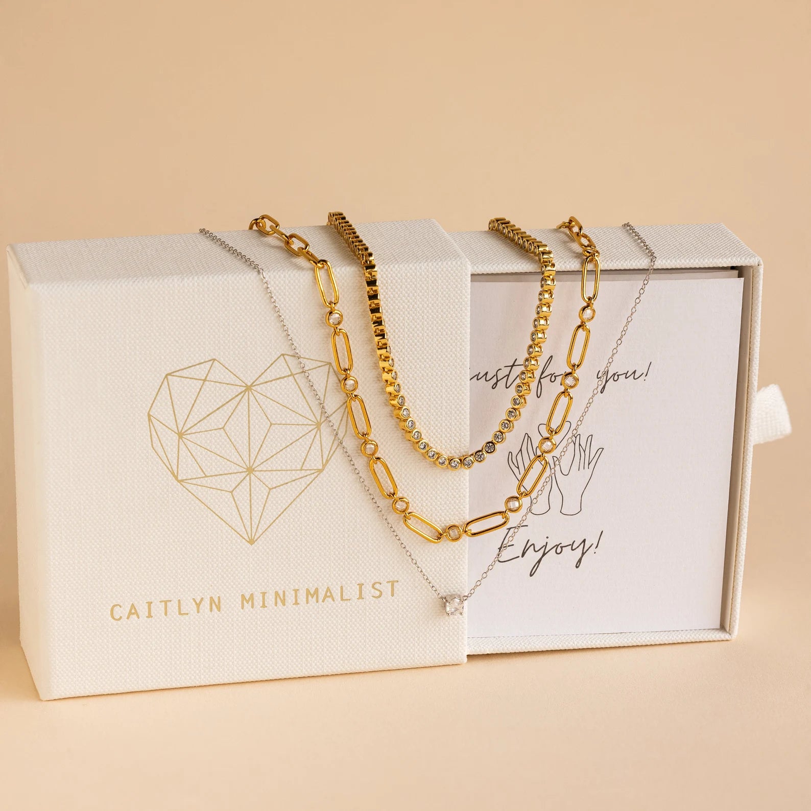 The Mystery Necklace Box features three layered gold and silver necklaces, elegantly presented on a cream Caitlyn Minimalist jewelry box.