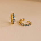 The Dainty Pave Birthstone Huggies are small gold hoop earrings with blue gemstones, displayed on a beige surface—perfect for adding everyday sparkle.