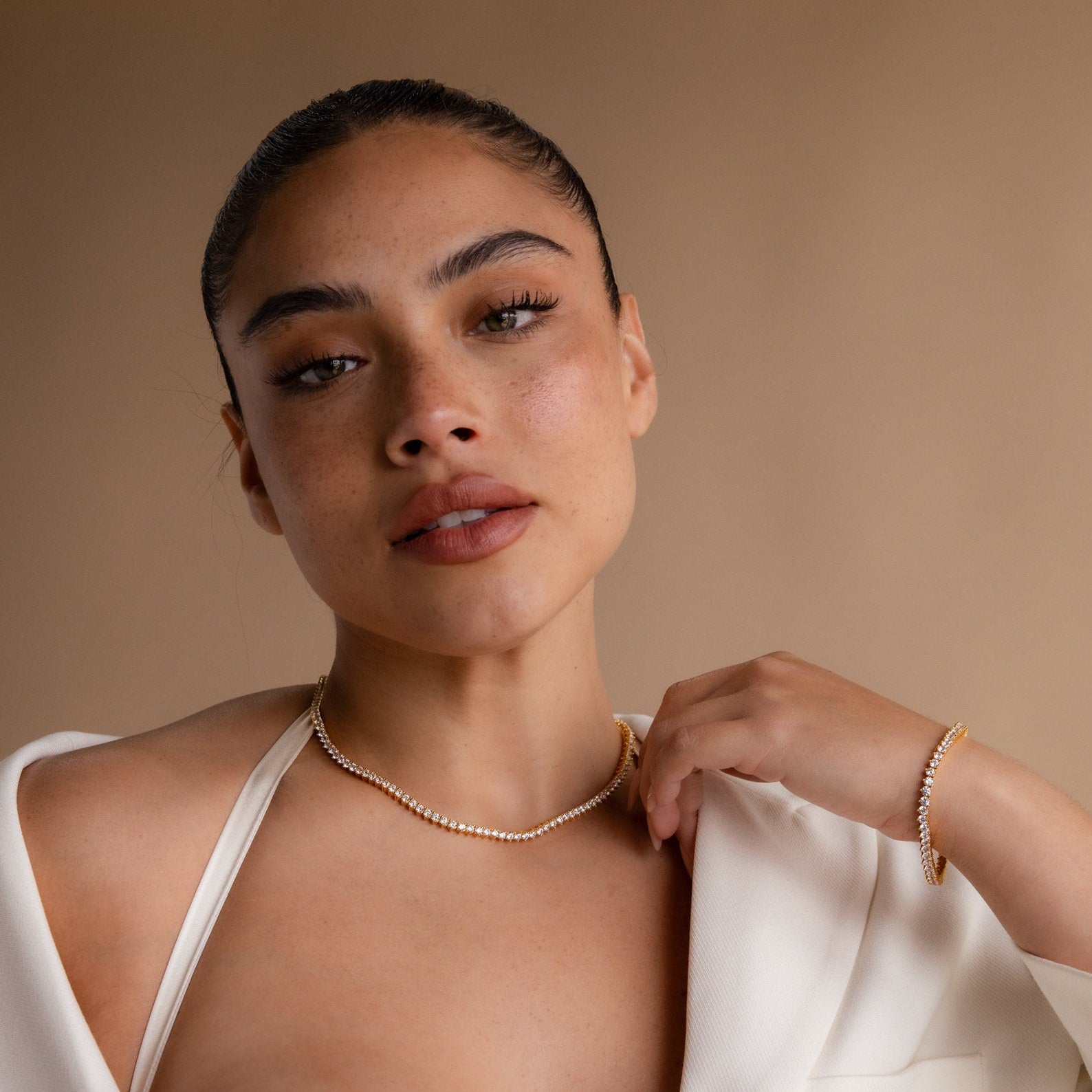 A woman in a white outfit poses gracefully against a beige background wearing the 3-Prong Diamond Tennis Necklace & Bracelet Set.
