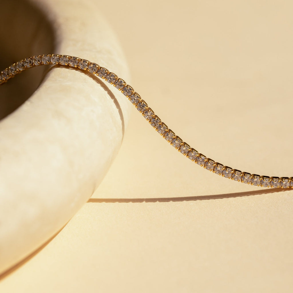 The Dainty Diamond Necklace Set features a gold tennis choker adorned with small round diamonds, elegantly draped over a smooth, light stone surface against a beige background.