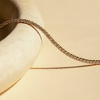 The Dainty Diamond Necklace Set features a gold tennis choker adorned with small round diamonds, elegantly draped over a smooth, light stone surface against a beige background.