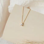 The Dainty Diamond Necklace Set features a gold chain with a single teardrop-shaped clear gemstone pendant, elegantly displayed on a light beige curved surface.