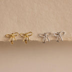 Two pairs of Classic Ribbon Bow Huggies in gold and silver are displayed on a textured beige surface, adding a touch of feminine elegance.