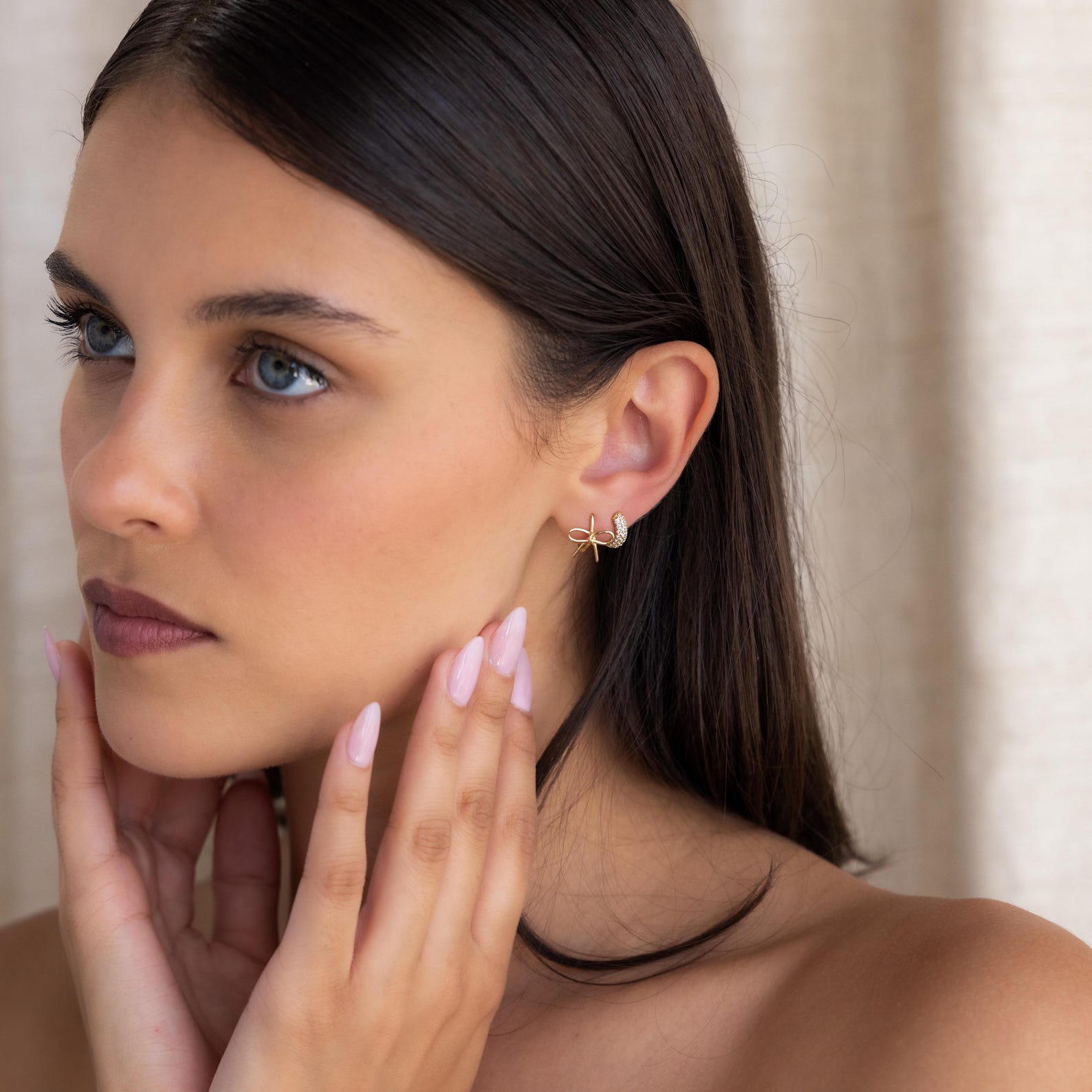 A woman with long dark hair touches her face, showing off elegant Classic Ribbon Bow Huggies earrings and pale pink nail polish.