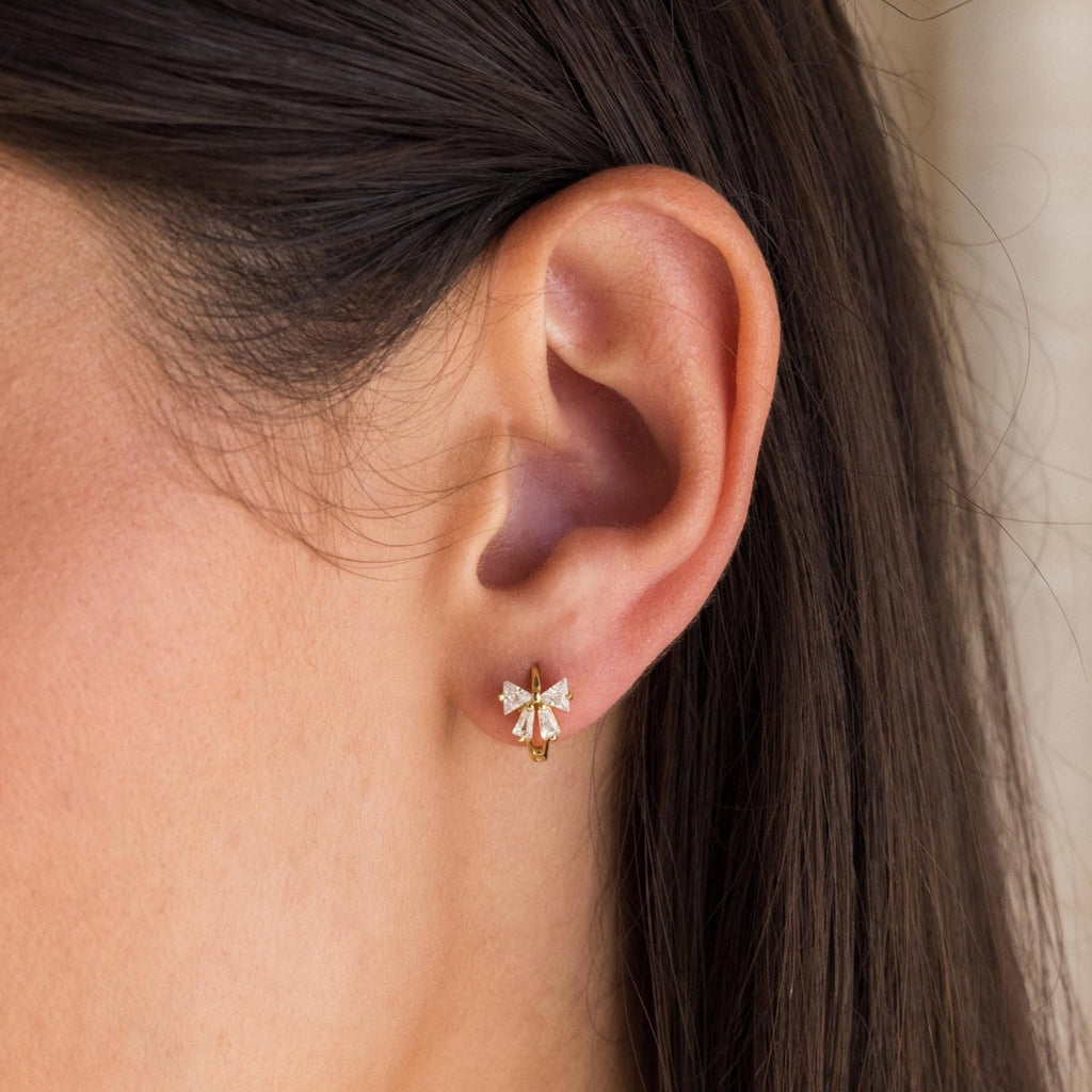 Close-up of a woman's ear wearing Diamond Ribbon Bow Huggies, featuring a small gold hoop with a decorative bow design.