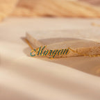 The Elegant Birthstone Name Necklace features “Morgan” in cursive with green stones on a beige surface—a stunning piece of personalized custom jewelry.