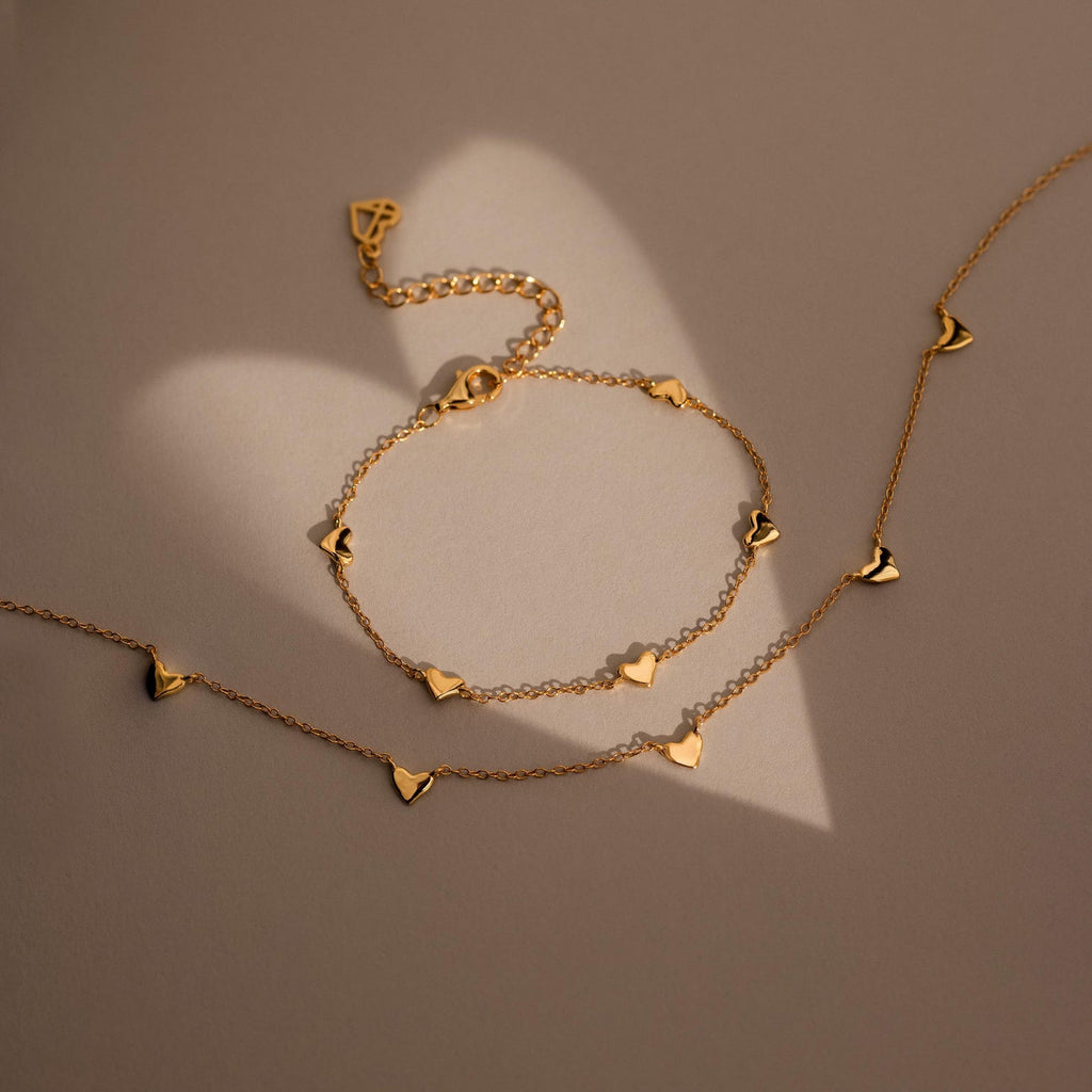 The Heart Station Necklace & Bracelet Set, featuring delicate charms, is arranged on a beige surface casting a heart-shaped shadow.