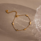 The Heart Station Necklace & Bracelet Set in 18K Gold features delicate heart charms, shown resting on a beige surface and softly illuminated.