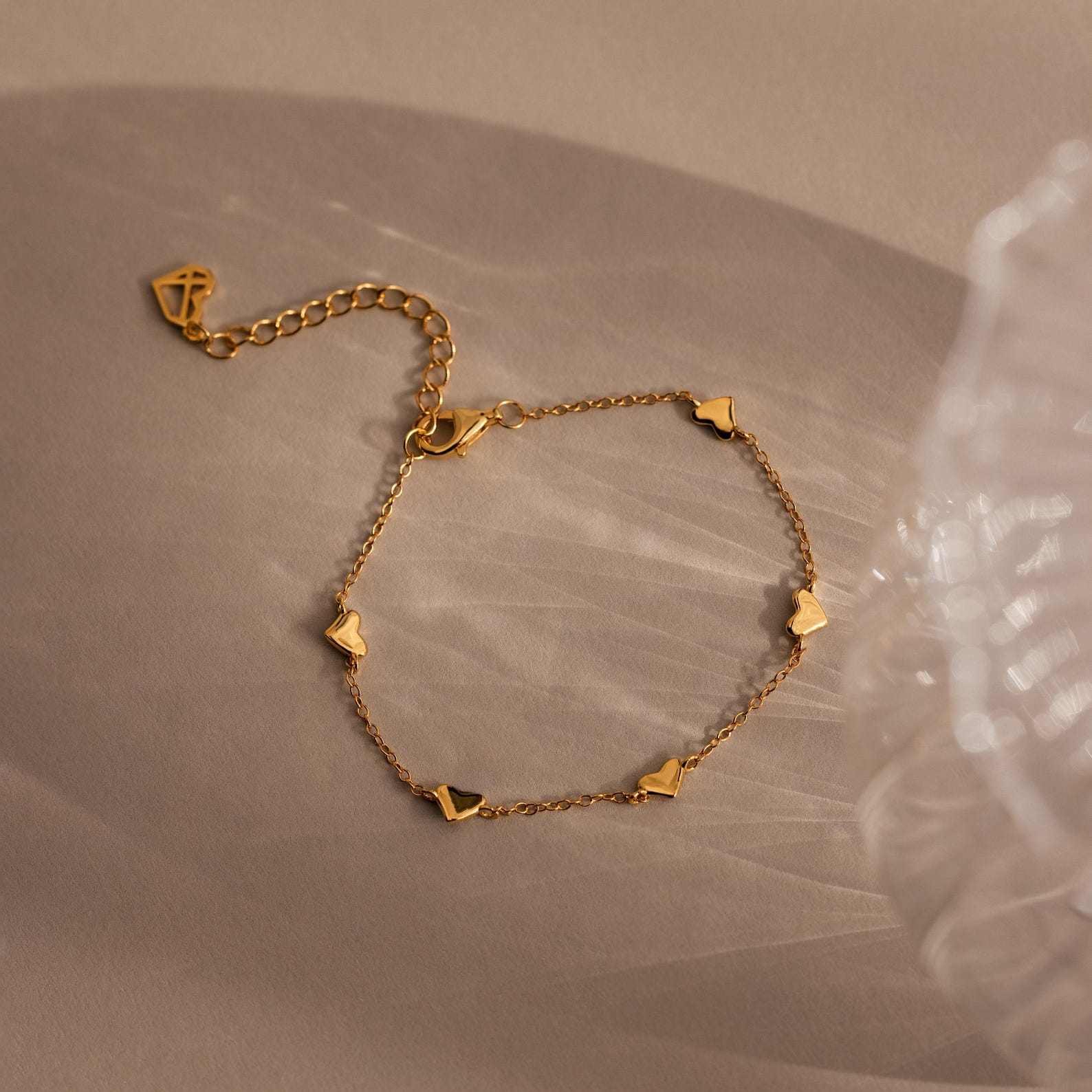 The Heart Station Necklace & Bracelet Set in 18K Gold features delicate heart charms, shown resting on a beige surface and softly illuminated.