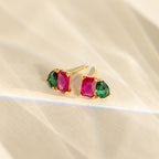 The Toi et Moi Birthstone Earrings feature rectangular pink and teardrop green stones set in gold, elegantly displayed on a textured surface. These vibrant earrings add a touch of color to any outfit.