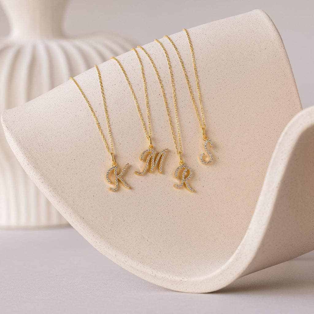 Four Large Pave Initial Necklaces with gold initial charms—K, M, R, and S—each accented with sparkling diamonds, are displayed on a curved beige surface.