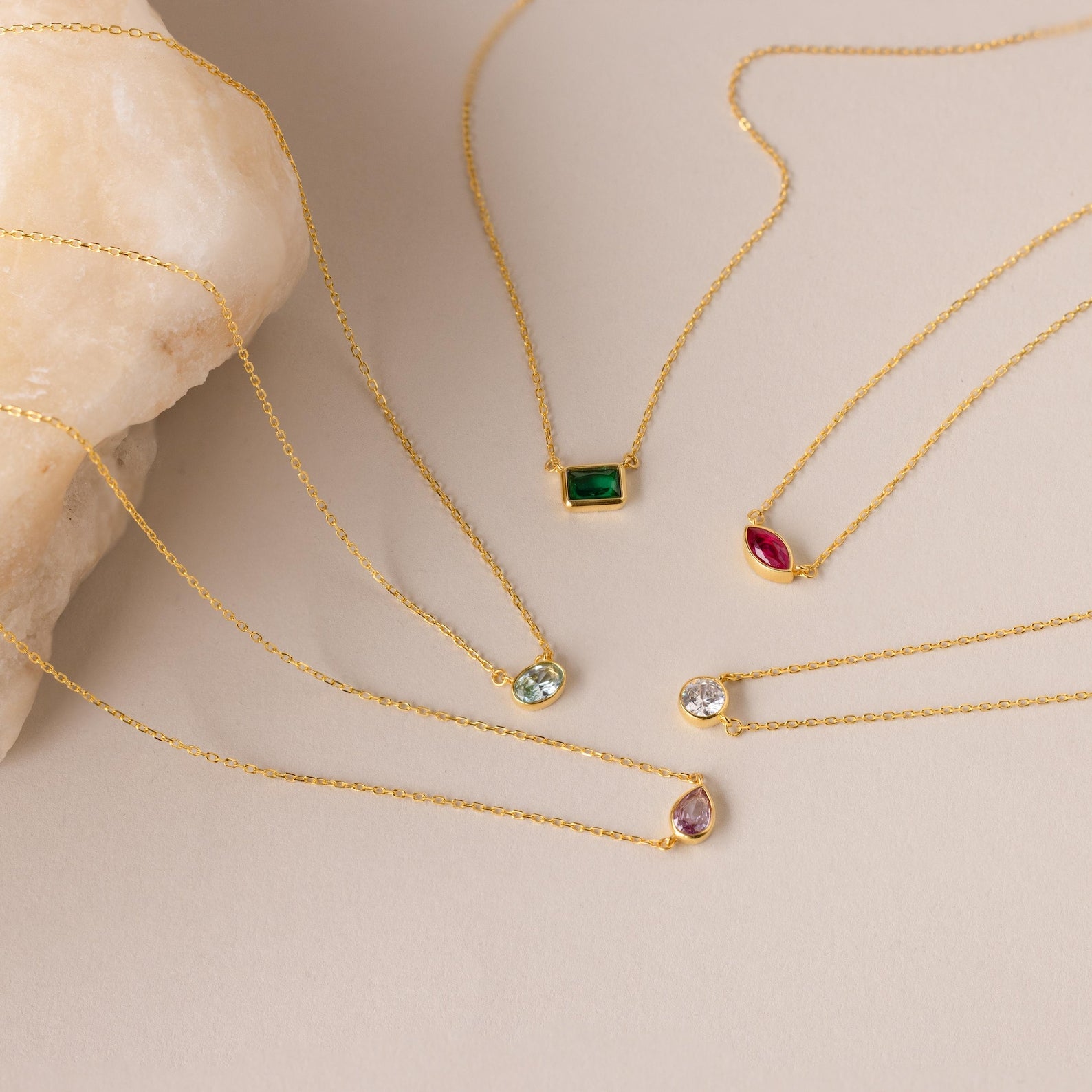 Five gold chain necklaces—including the Custom Cut Birthstone Necklace—are displayed on a beige surface beside a stone, making them ideal for fans of personalized jewelry.