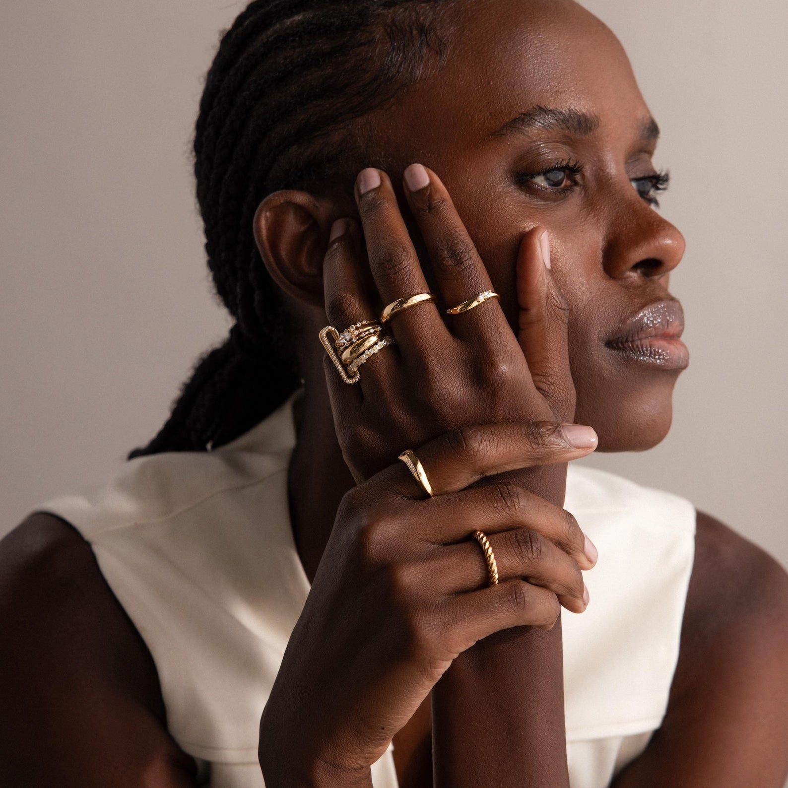 A woman with cornrows, wearing multiple gold rings including the Multipurpose Pave Link Clasp Set in 18K Gold, rests her face on her hand, thoughtfully showcasing layered jewelry styling.