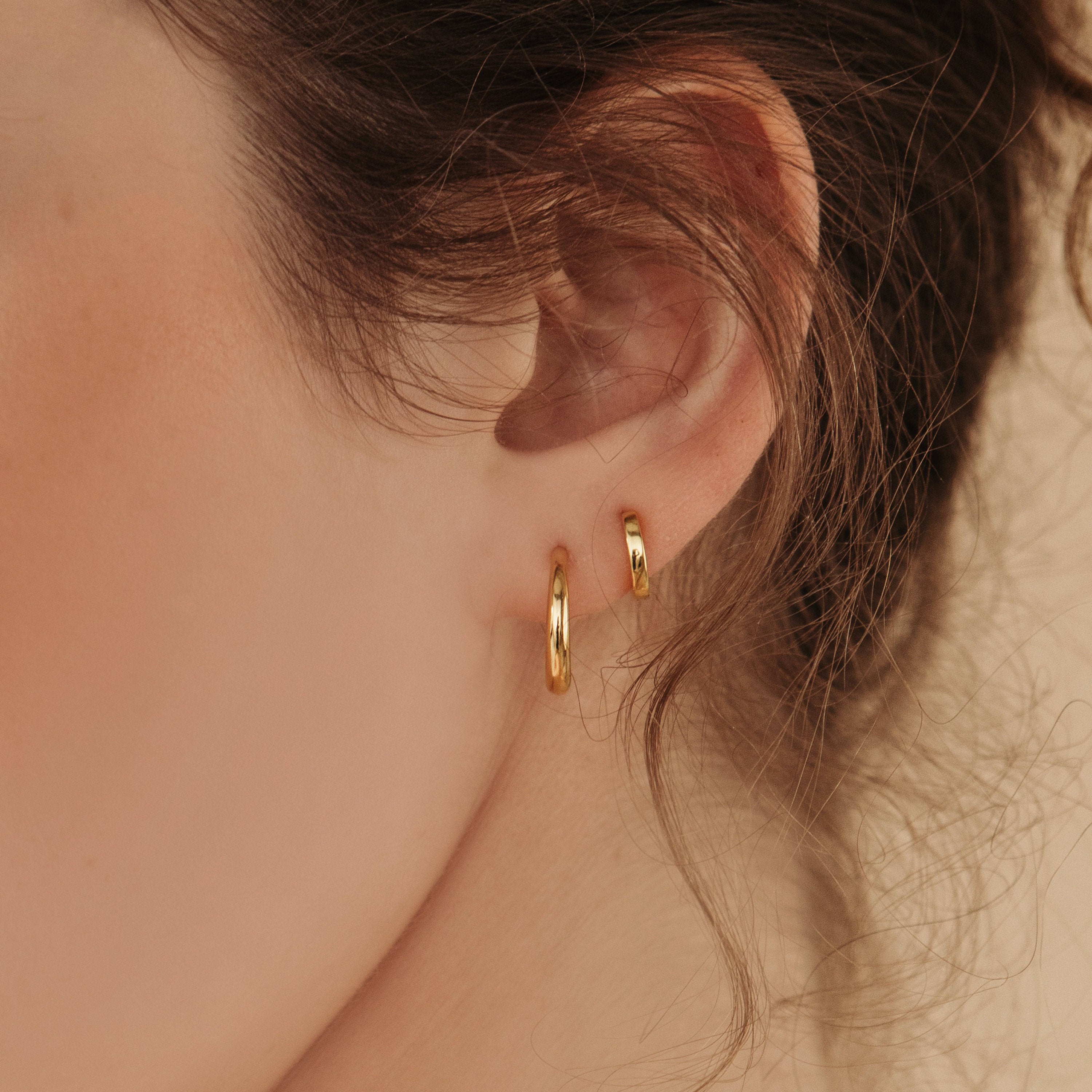Close-up of a woman's ear with two Editor Huggies in gold and wavy brown hair, highlighting a minimal aesthetic.