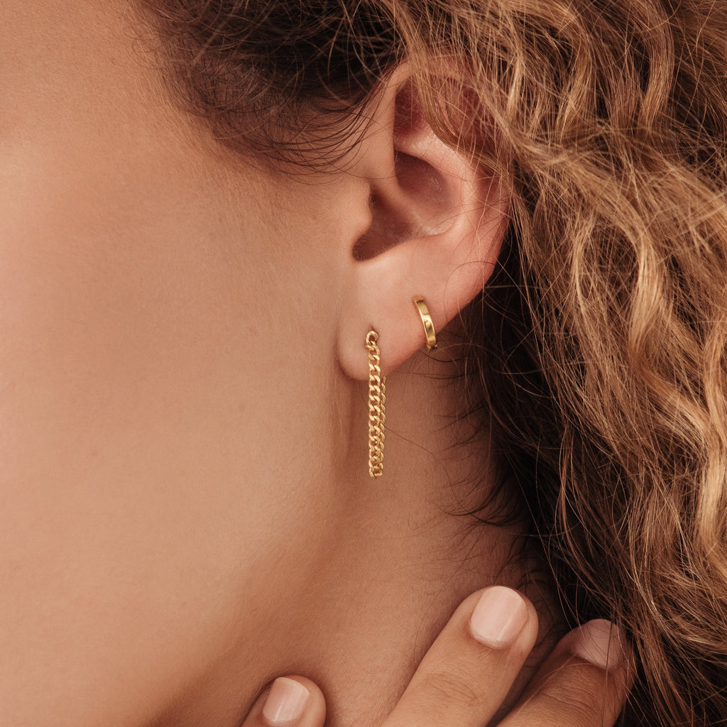 Close-up of a woman's ear wearing Editor Huggies in Rose Gold—one small hoop and one with a delicate hanging chain—highlighting minimal, classic jewelry style.