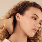 A woman with curly hair wears Editor Huggies in 18K Gold and a gold bracelet, her hand resting on her head against a neutral background.