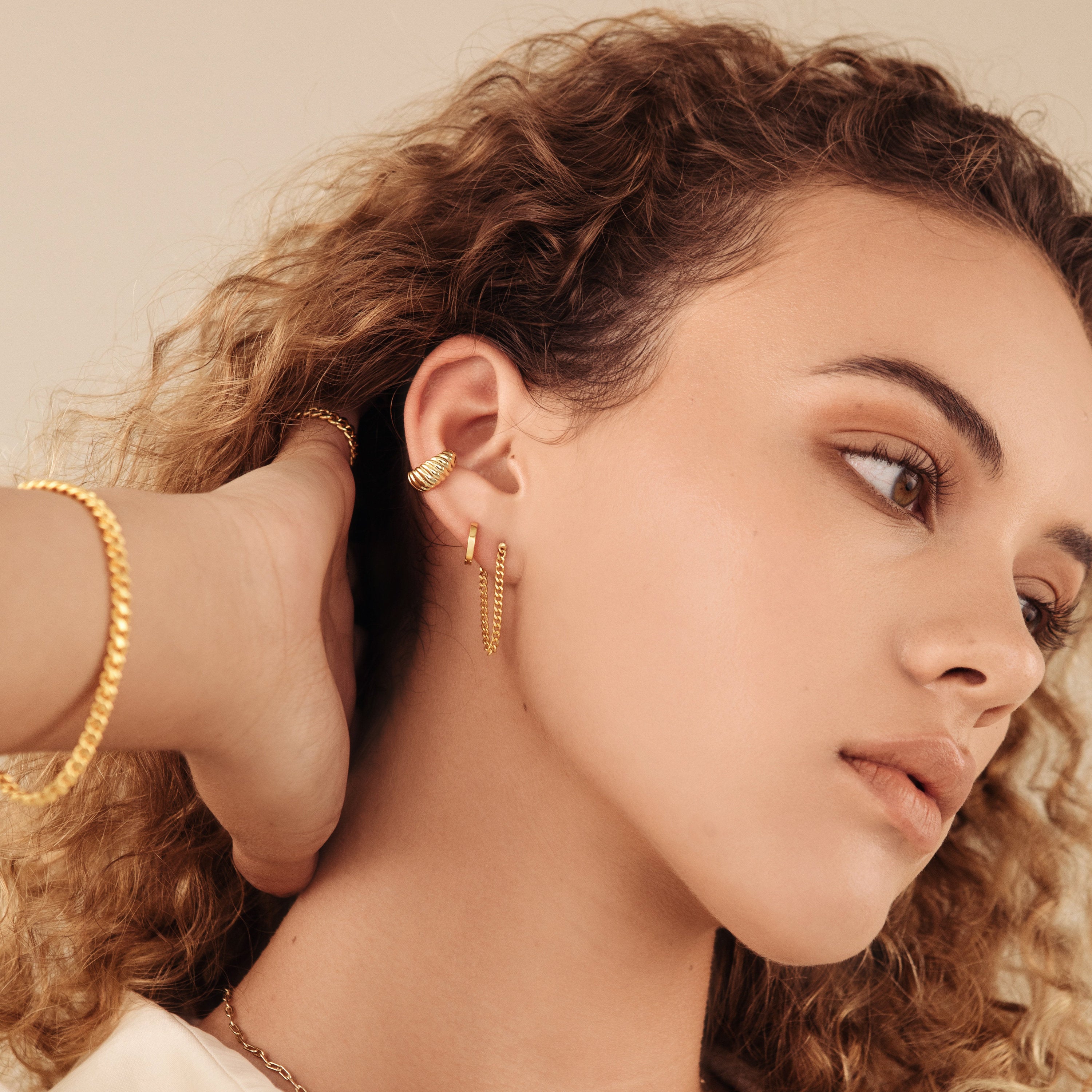 A woman with curly hair wears classic jewelry—Editor Huggies in rose gold and a gold bracelet—as she poses with her hand behind her head.
