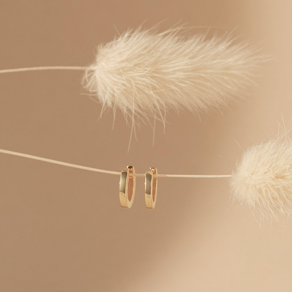 Editor Huggies in Rose Gold hang on a string between two fluffy dried plants against a beige background, highlighting their minimal aesthetic.