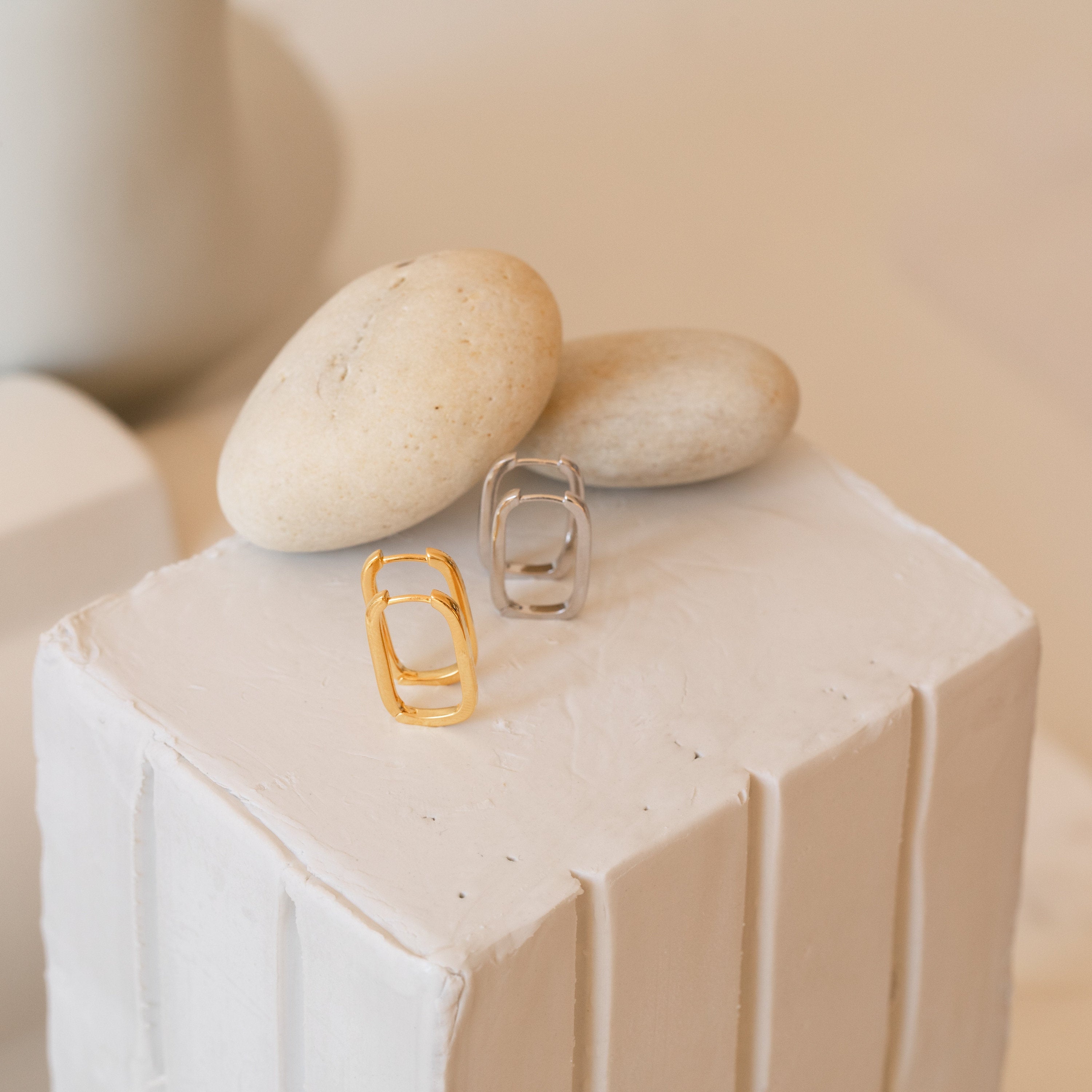 The Linked Earrings Set, featuring gold and silver modern designs, is displayed on a white textured surface with smooth beige stones—perfect for enhancing your jewelry collection.