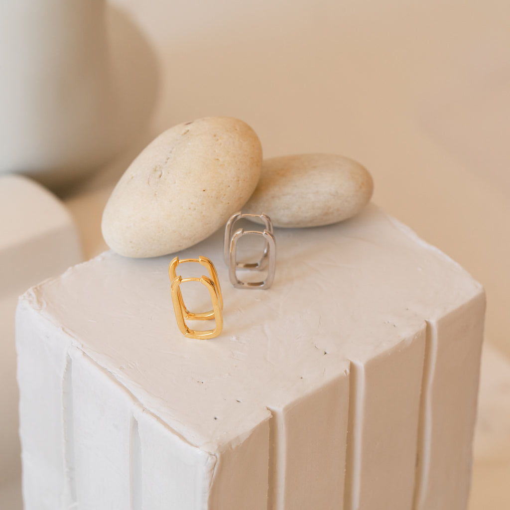 Nina Link Earrings in gold and silver are shown on a white textured platform with smooth beige stones, emphasizing the modern style of these geometric hoops.
