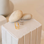 Nina Link Earrings in gold and silver are shown on a white textured platform with smooth beige stones, emphasizing the modern style of these geometric hoops.