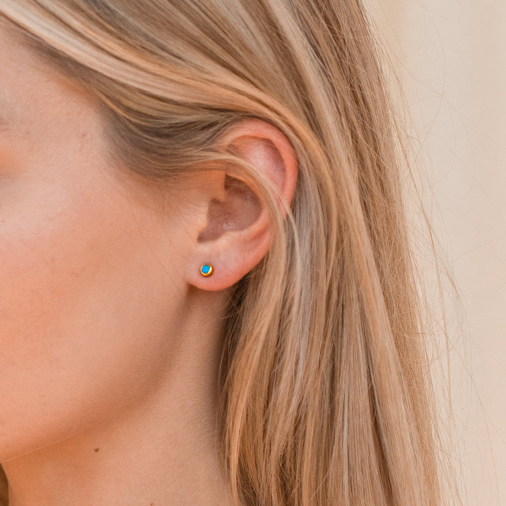 Close-up of a model wearing a small stud earring with a colorful gold turquoise gemstone, against a neutral background. 