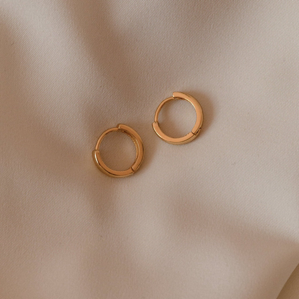 Two Editor II Huggies in sterling silver rest on a beige fabric background, perfect for everyday wear or adding subtle elegance—these earrings are a timeless essential.