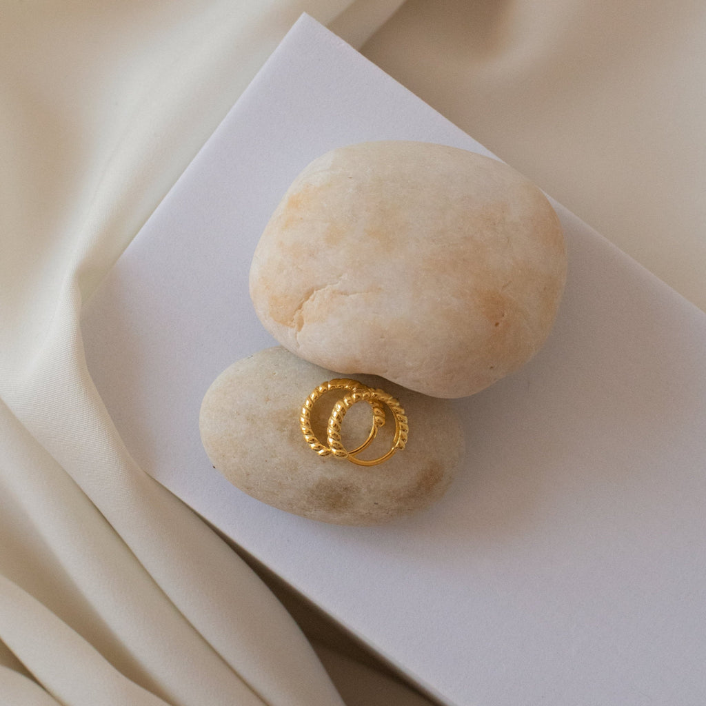 Thea Twisted Huggies rest on smooth beige stones atop a white surface with draped cream fabric—a perfect minimalist choice for your earrings or ear stack collection.