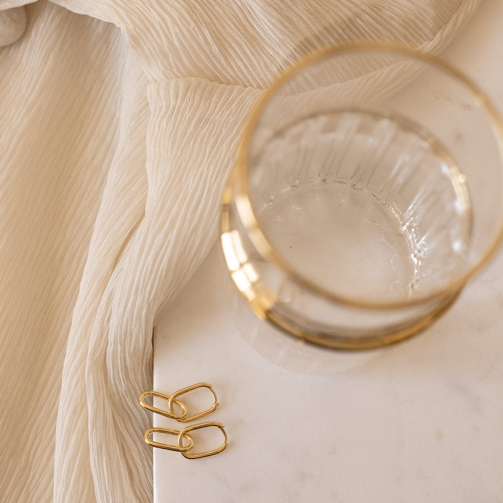 The Linked Earrings Set from our jewelry collection sits on a marble surface paired with cream textured fabric and gold-rimmed glass.