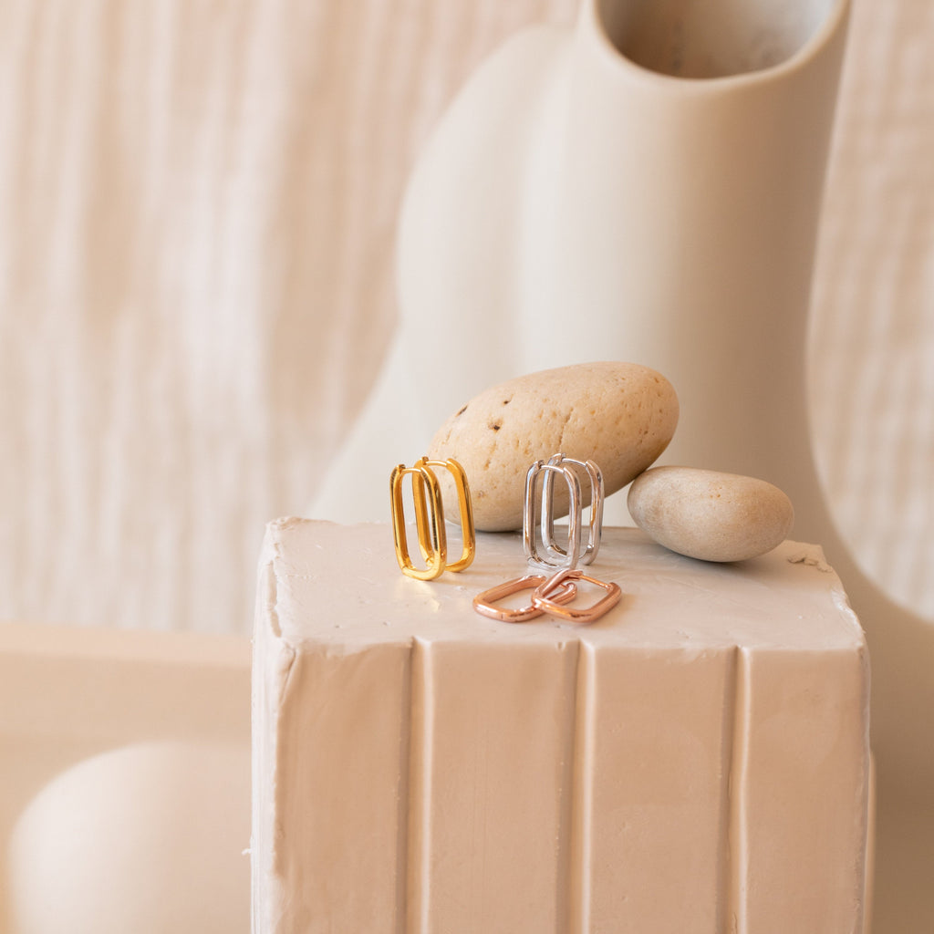 Three pairs of Nina Link Earrings—geometric metal hoops in gold, silver, and rose gold—are displayed on a white textured pedestal with stones.