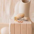 Three pairs of Nina Link Earrings—geometric metal hoops in gold, silver, and rose gold—are displayed on a white textured pedestal with stones.